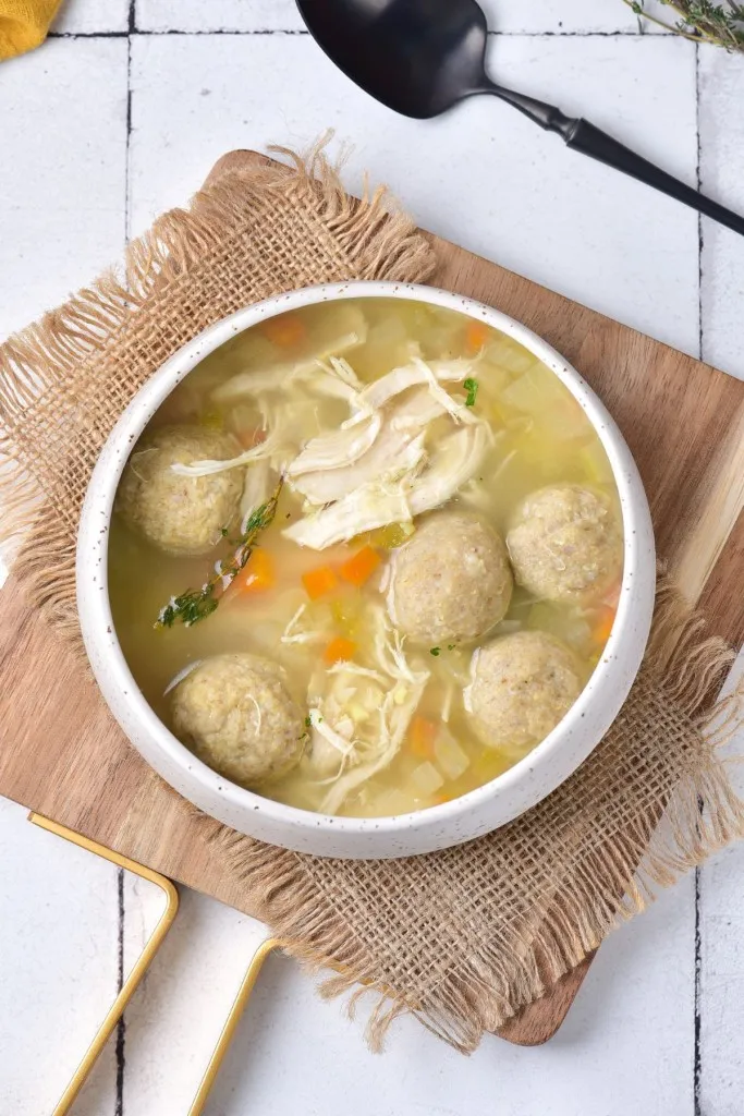 Overhead view of a bowl of Matzo ball soup on a wooden kitchen board and a black serving spoon on a white countertop.