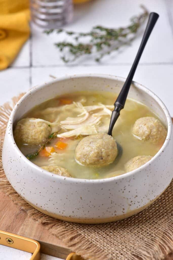 Bowl of Matzo ball soup with black spoon on a wooden kitchen board.