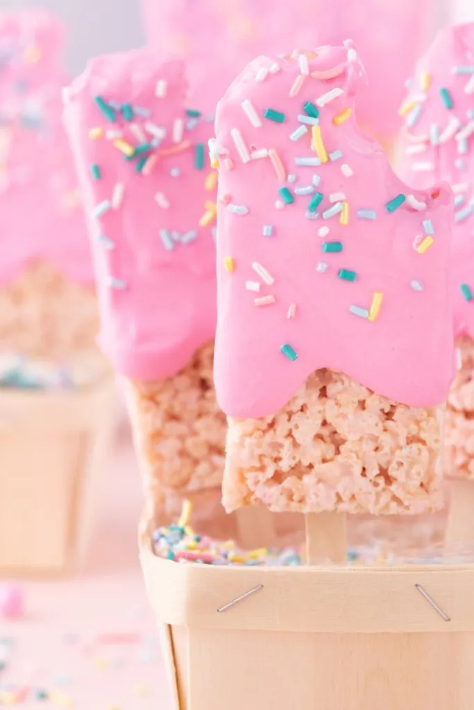 Close up of Pink Popsicle Rice Krispie Treats standing upright in a container on a pink surface.