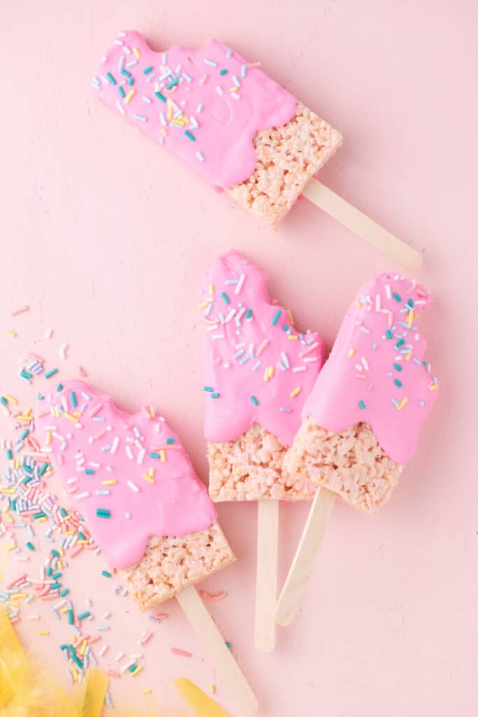 Four Pink Popsicle Rice Krispie Treats on a pink surface with colored sprinkles.