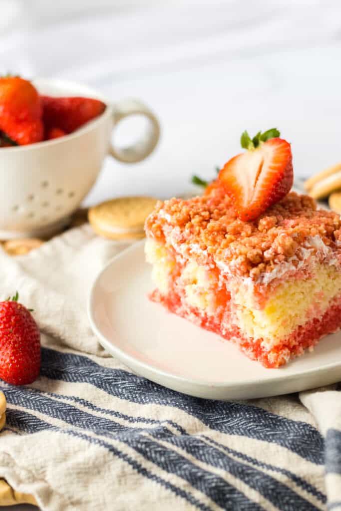 Slice of Strawberry Crunch Poke Cake on a white plate, tea cup with fresh strawberries, blue and cream striped kitchen cloth on a white countertop.