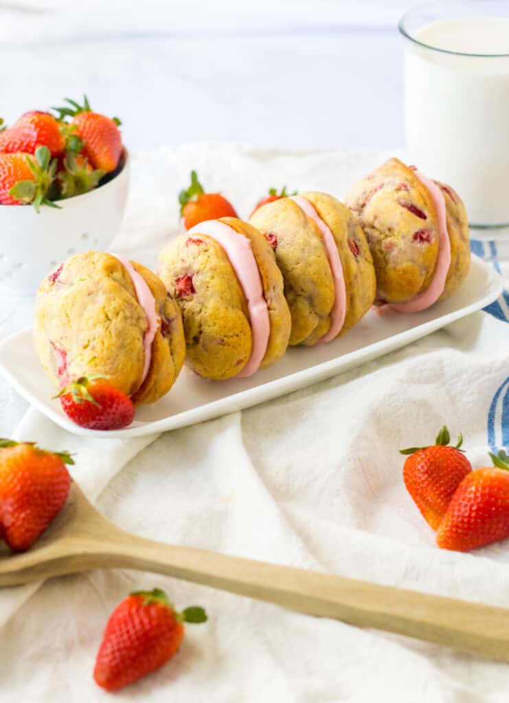 Four assembled Strawberry Shortcake Whoopie Pies on a white serving plate, bowl of fresh strawberries, glass of milk, blue and cream striped kitchen towel.