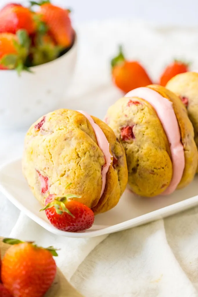 Close up of Strawberry Shortcake Whoopie Pies on a white platter, bowl of fresh strawberries, blue and cream striped kitchen towel.