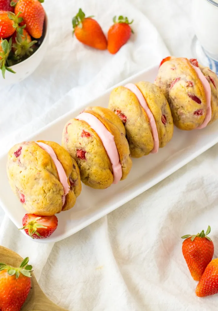 Overhead view of Strawberry Shortcake Whoopie Pies on a white platter, fresh strawberries, on a blue and cream striped kitchen towel.