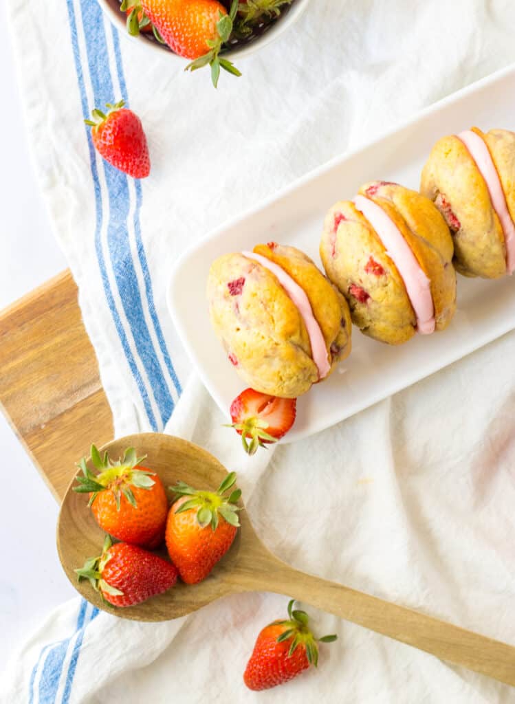 Overhead view of Strawberry Shortcake Whoopie Pies on a white plate, wooden spoon with fresh strawberries, bowl with fresh strawberries, blue and cream striped kitchen towel on a wooden kitchen board.