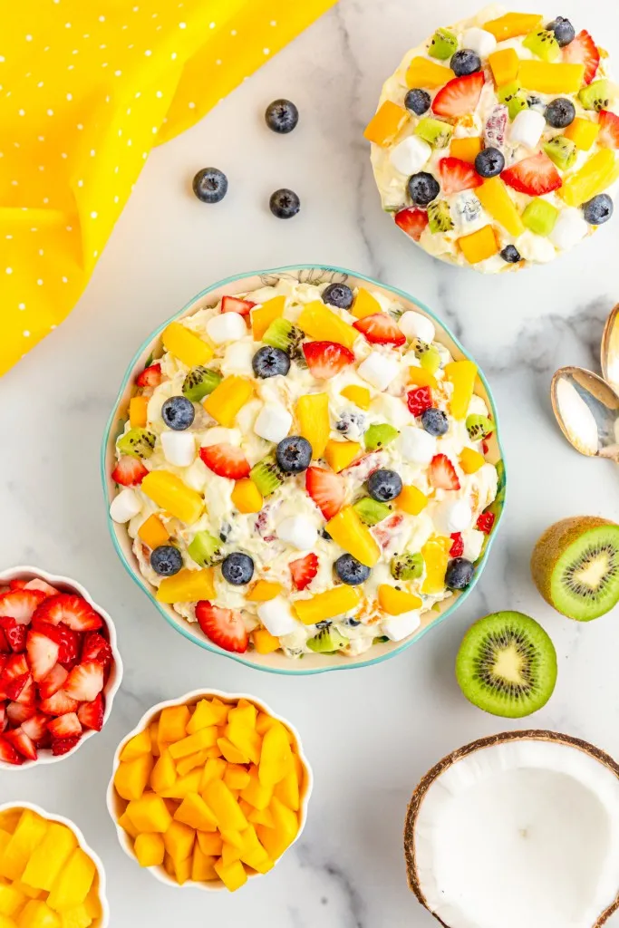Overhead view of tropical fruit marshmallow fluff, fresh fruit, yellow kitchen cloth on a white countertop.
