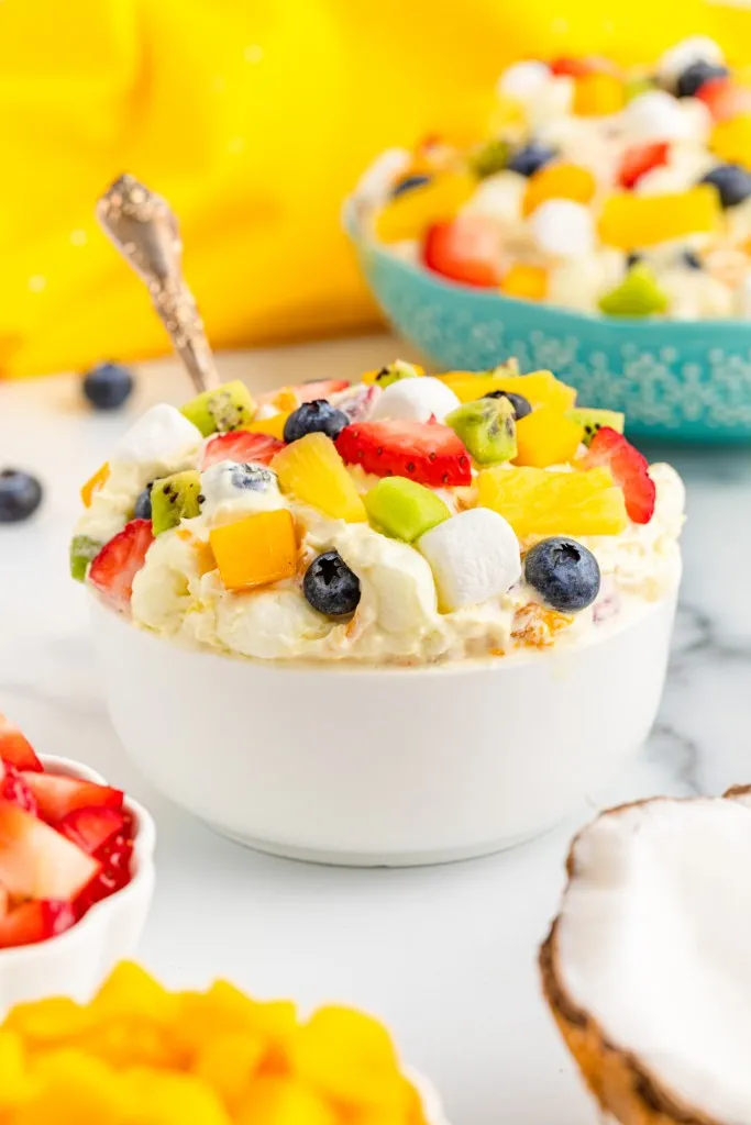 Single serving bowl of tropical fruit marshmallow fluff, fresh fruit, larger bowl of fluff dessert, yellow kitchen cloth on a white countertop.