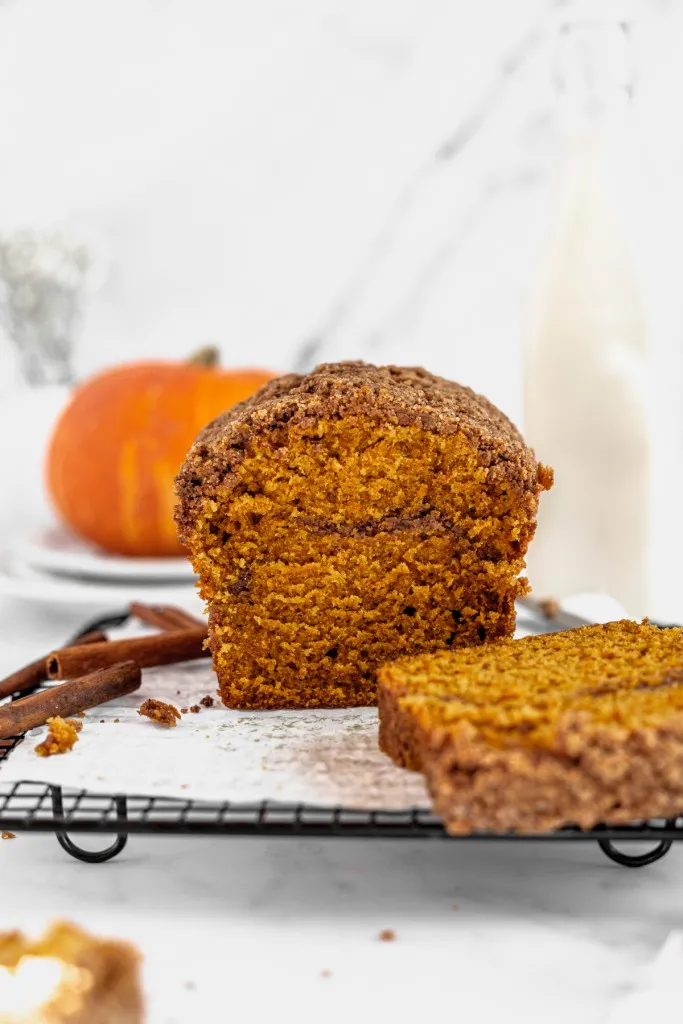 Pumpkin streusel bread loaf on a wire cooling rack, whole pumpkin and loose cinnamon sticks on a white countertop.