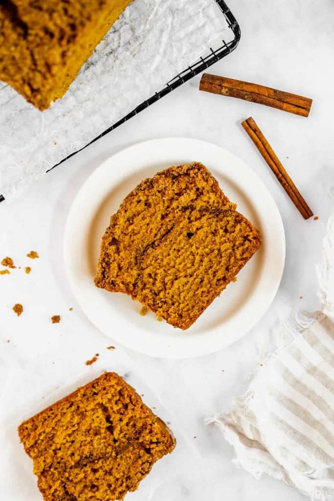 Overhead view slices of pumpkin streusel bread, pumpkin streusel bread loaf on a wire rack, loose cinnamon sticks and kitchen cloth on a white countertop. 