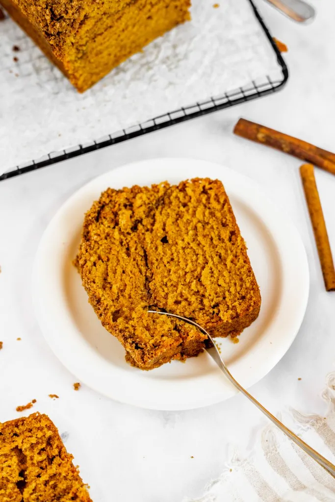 Slice of pumpkin streusel bread on a white plate with metal fork, pumpkin streusel bread loaf on a wire cooling rack, on a white countertop.