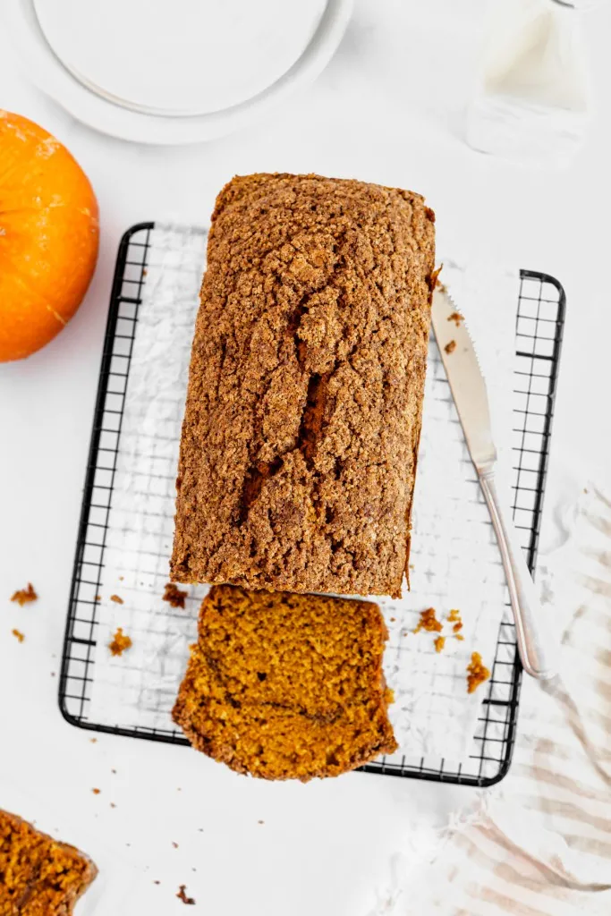 Overhead view of pumpkin streusel bread on a wire cooling rack with metal bread knife, whole pumpkin and other recipe ingredients surrounding the bread.