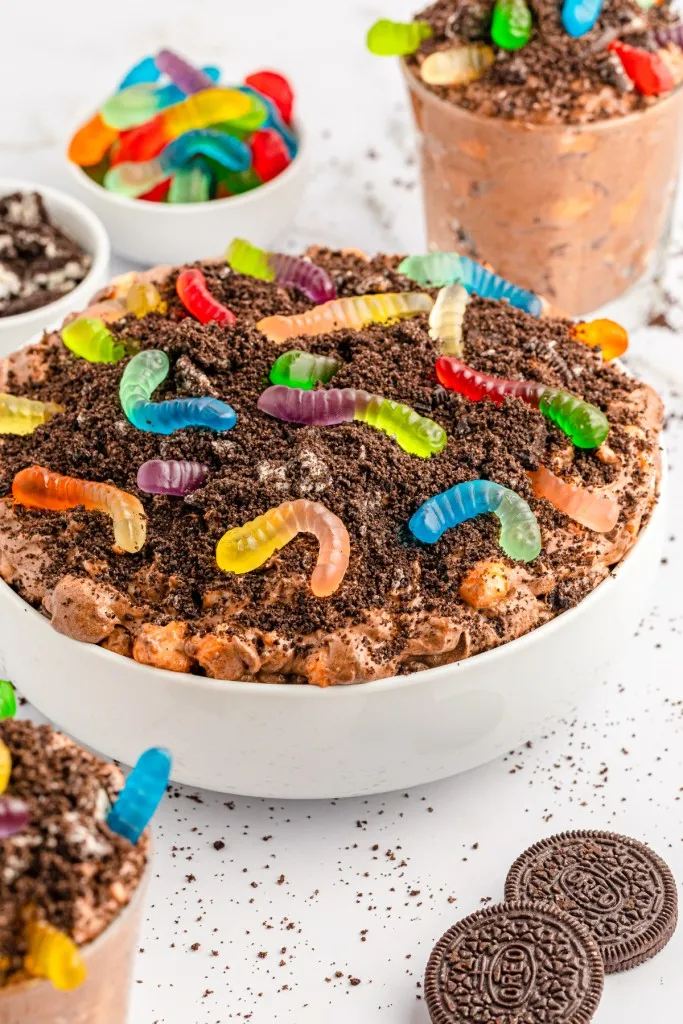 Bowl of dirt worm pudding topped with crushed Oreos and gummy worms, dirt worm cups, loose Oreos, small bowls of gummy worms and broken Oreos, on a white countertop.