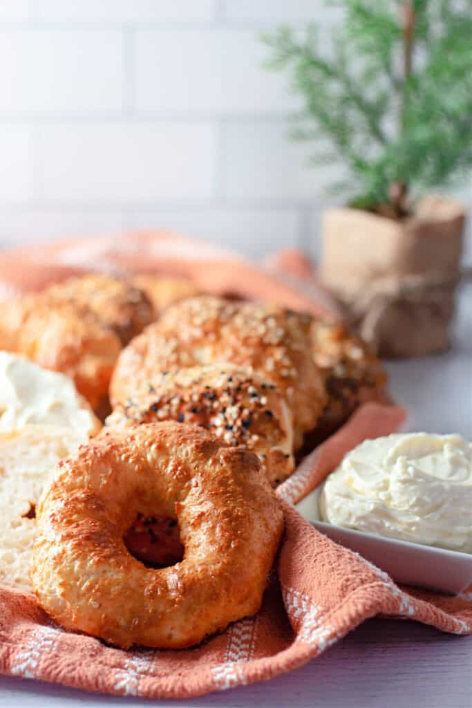 Row of air fryer bagels on a kitchen cloth with a bowl of cream cheese spread on a white countertop.