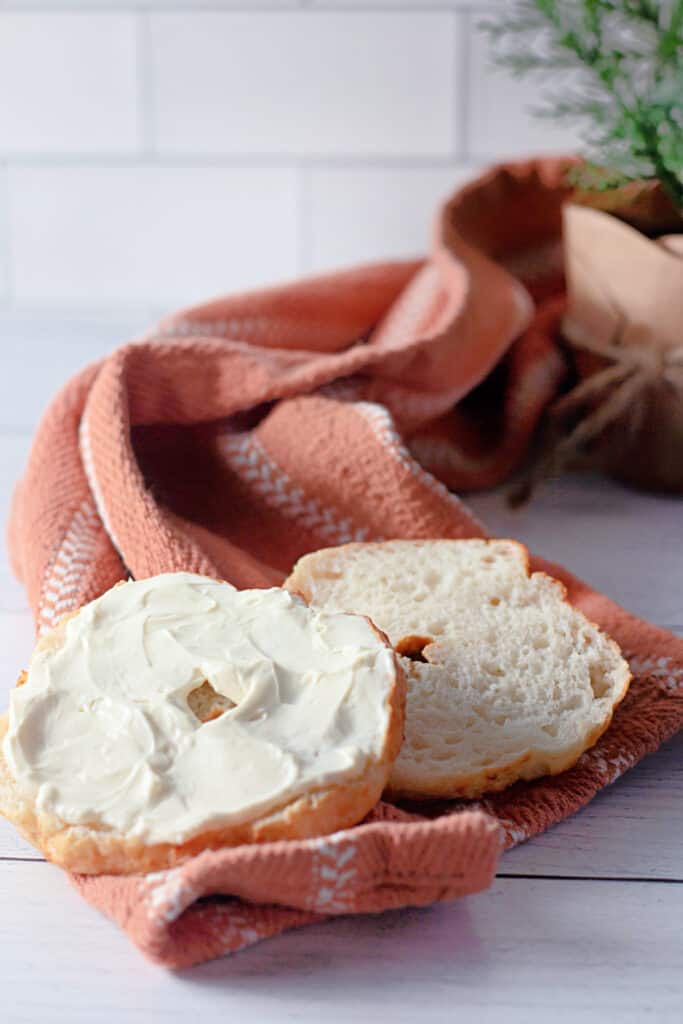 Sliced air fryer bagel with cream cheese spread on a kitchen cloth on a white countertop.