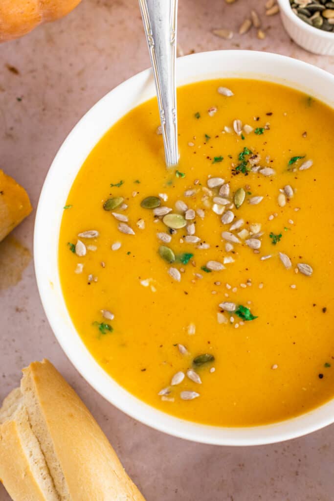 Bowl of easy pumpkin soup recipe with a metal spoon, bread roll and recipe ingredients on the countertop topped with sunflower seeds and pepitas.