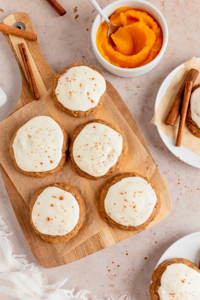 Overhead view of iced pumpkin cookies on a wooden kitchen board, pumpkin cookie ingredients surrounding the cookies, on a countertop.