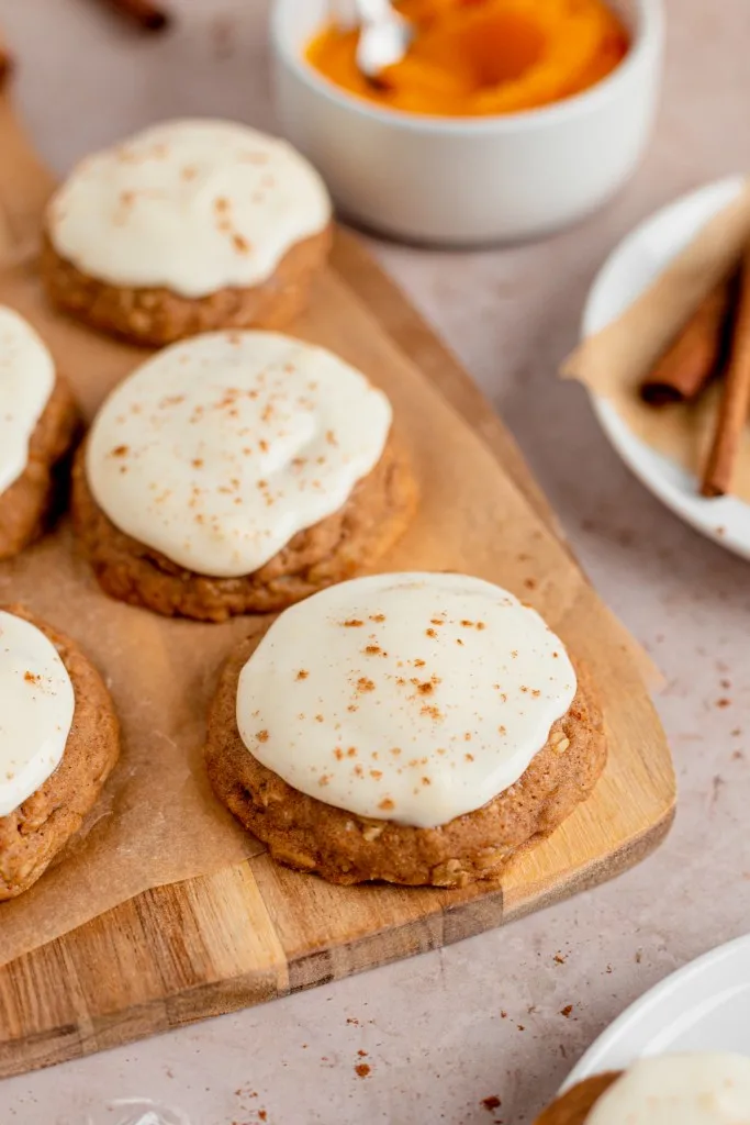 Iced pumpkin cookies on a wooden kitchen board with recipe ingredients surrounding the cookies.