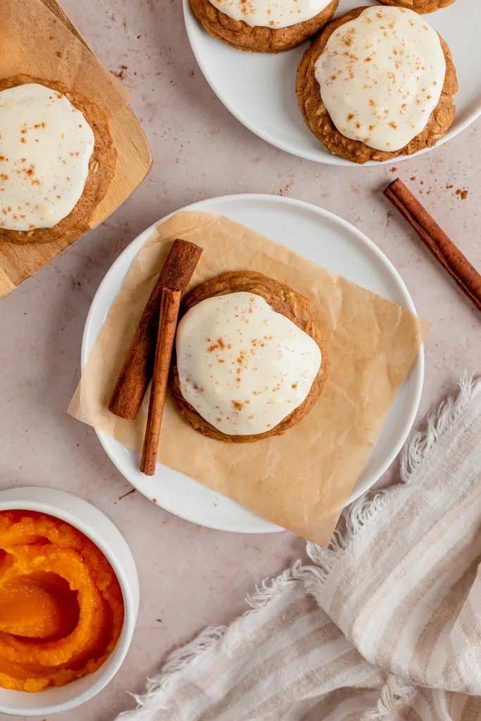 Overhead view of iced pumpkin cookies, bowl of pumpkin puree, loose cinnamon sticks, kitchen cloth.