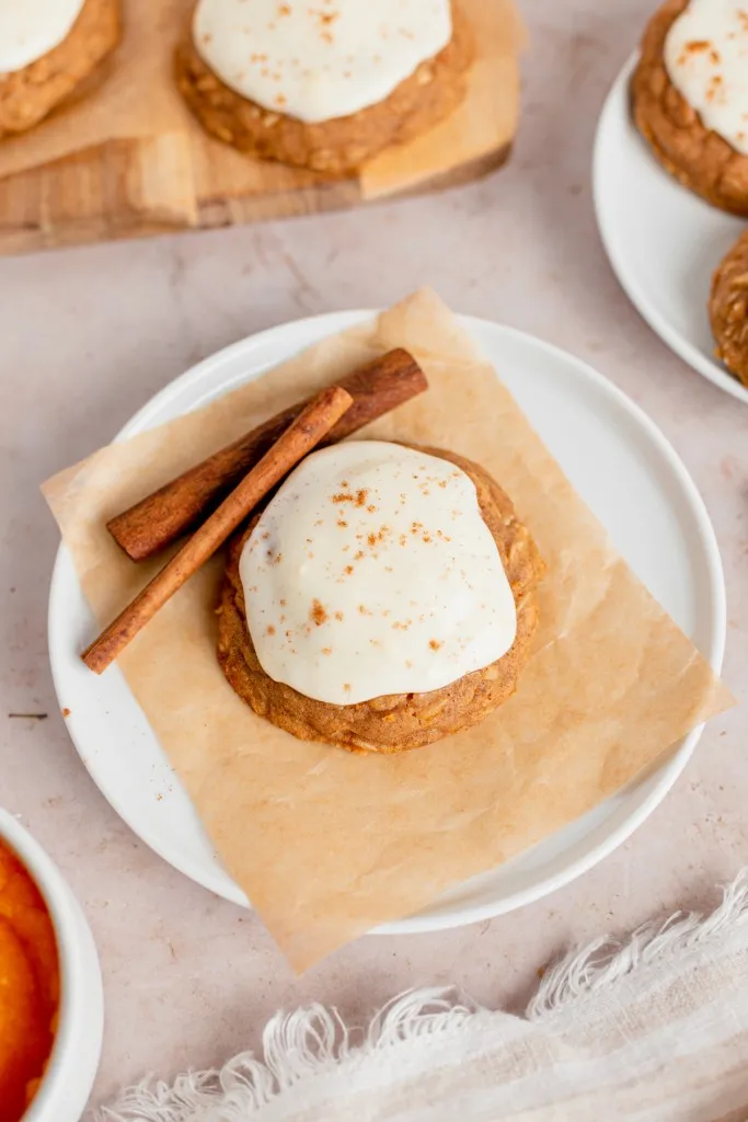 Overhead view of an iced pumpkin cookie on a serving plate with loose cinnamon sticks, additional cookies in the background.