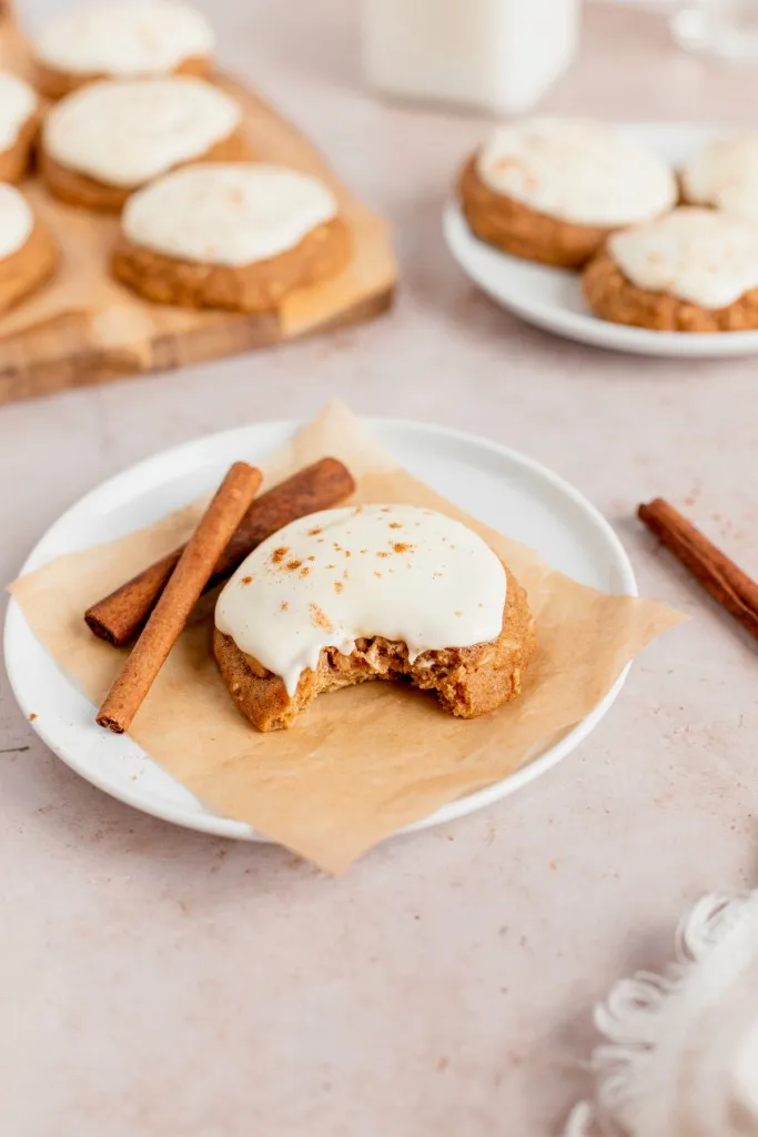 Iced Pumpkin cookies on a plate with a bite taken out of it, loose cinnamon sticks and remaining iced pumpkin cookies in the background.