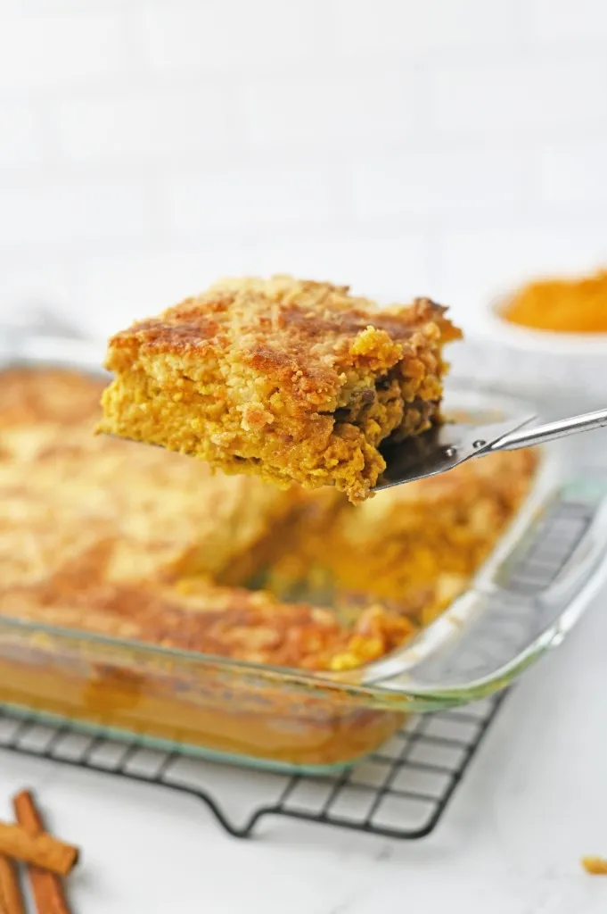 Silver server holding a slice of pumpkin dump cake, remaining pumpkin dump cake in a glass baking dish on a wire cooling wrack on a white kitchen countertop.
