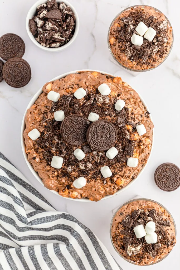 Overhead view of Oreo Fluff Salad in a large bowl and smaller bowls, striped kitchen cloth, small bowl of crushed Oreos, whole Oreos on a white marble countertop.