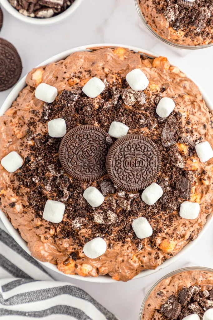 Overhead view of bowl of Oreo Fluff Salad, single glasses filled with Oreo Fluff Salad, striped kitchen towel, small bowl of crushed Oreos, whole Oreos on a marble countertop.