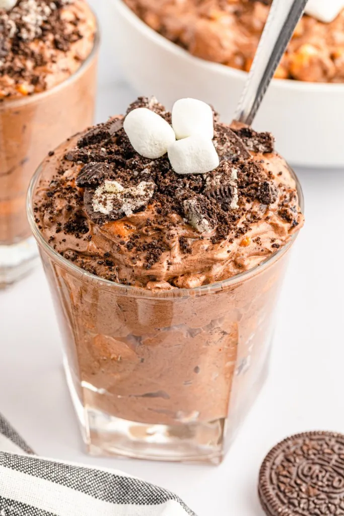 Oreo fluff salad in serving glasses with a metal spoon, bowl of Oreo Fluff Salad, striped kitchen cloth, whole Oreos on a white marble countertop.