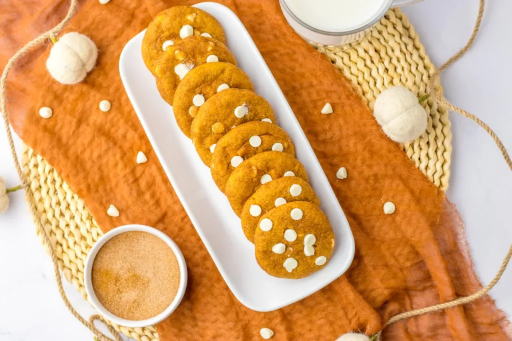 Overhead view of white serving tray with fall inspired snickerdoodle cookies, glass of milk, bowl of cinnamon sugar, fall-inspired table decoration.