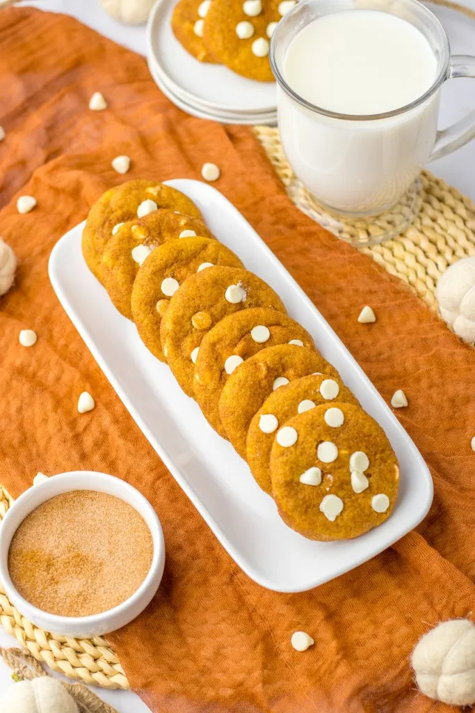 Overhead view of white serving tray with pumpkin snickerdoodle cookies, stacked white plates with pumpkin snickerdoodle cookies, glass of milk, bowl of cinnamon sugar, fall-inspired table decoration. 