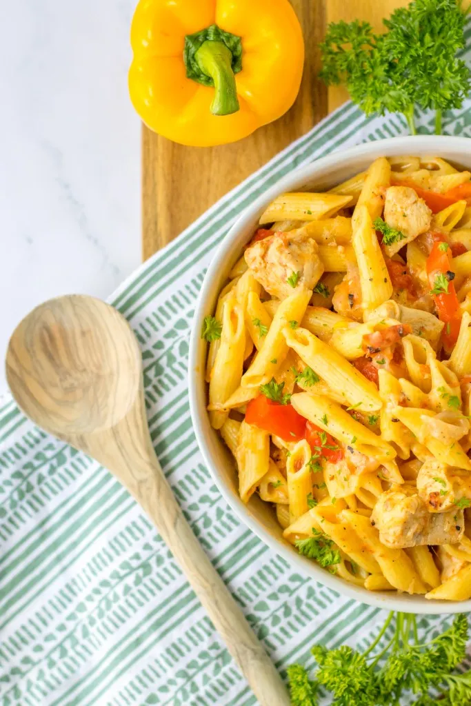 Overhead view of a bowl of slow cooker Cajun chicken pasta with wooden spoon, yellow bell pepper, fresh parsley, and decorative kitchen cloth.