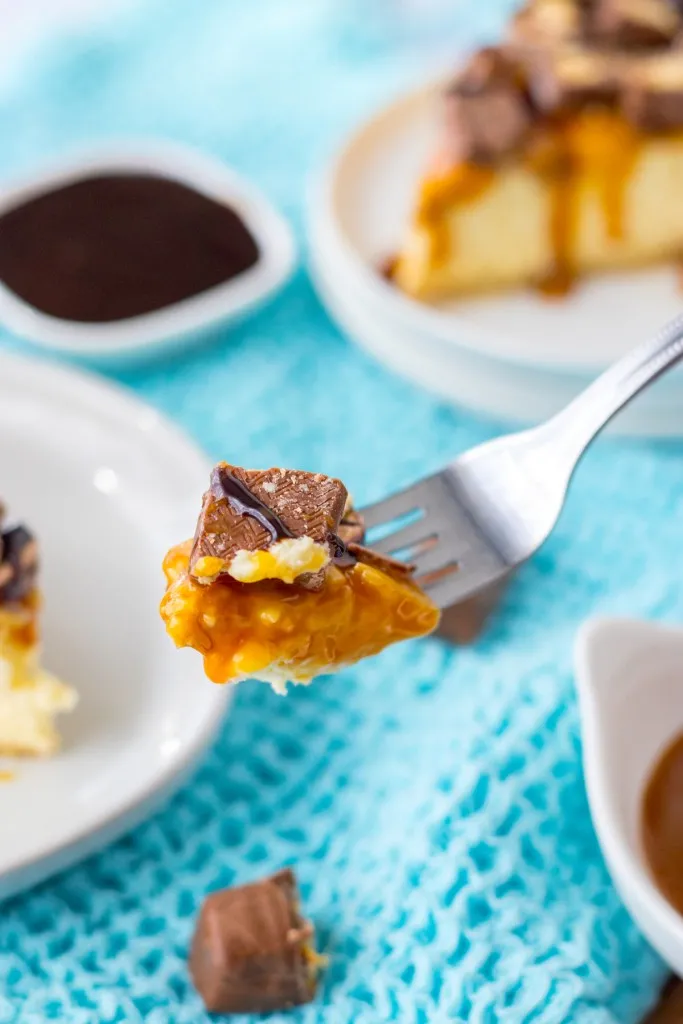 Fork holding a piece of Twix cheesecake, two white plates with slices of Twix cheesecake in the background with a small bowl of chocolate syrup, on a light blue kitchen cloth.