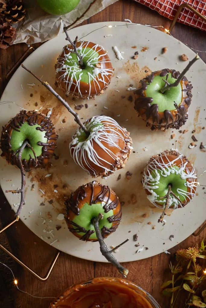 Overhead view of best caramel apples on a large serving board, bowl of caramel sauce, pine cones in the background.