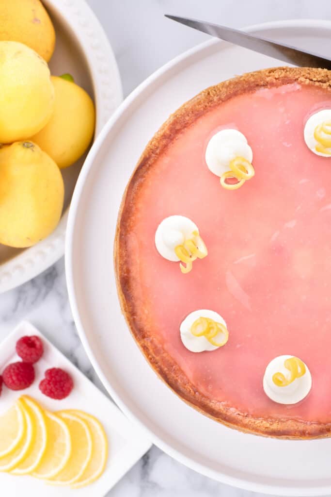 Lemon raspberry cheesecake on a white plate, whole lemons and raspberries on a marble countertop.
