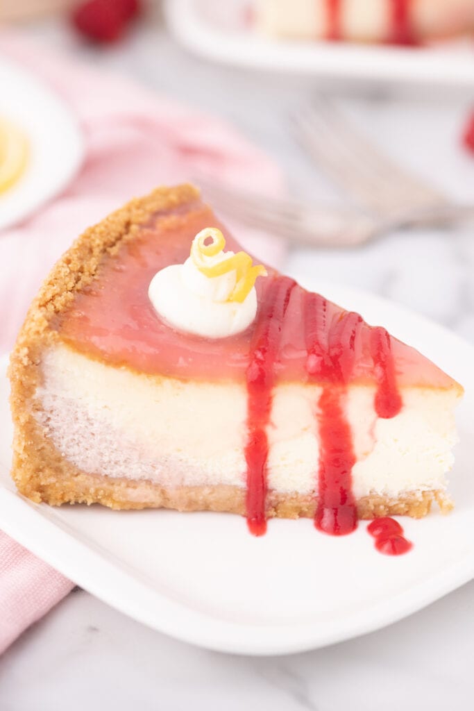 Slice of lemon raspberry cheesecake on a white plate, pink cloth and larger cheesecake in the background.