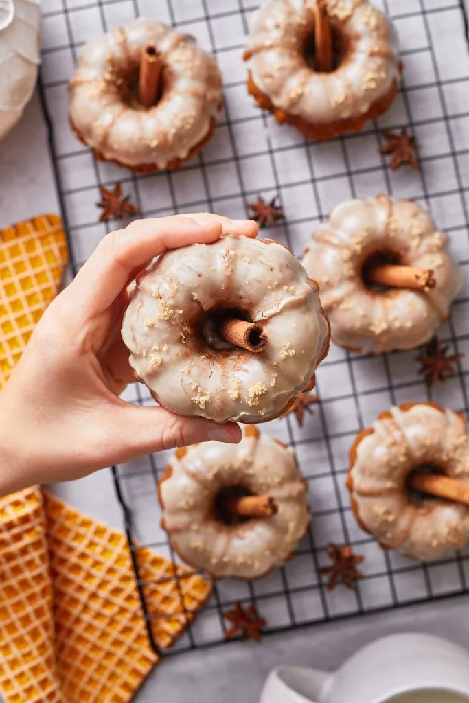 Hand holding a mini pumpkin bundt cake, mini pumpkin bundt cakes on a wire rack, orange cloth on a white countertop.