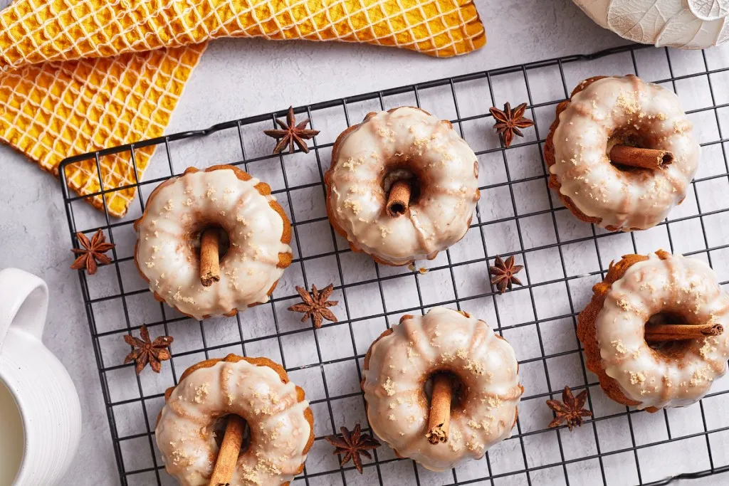 Mini pumpkin bundt cakes on a wire rack with cinnamon sticks, orange kitchen towel on a white countertop.