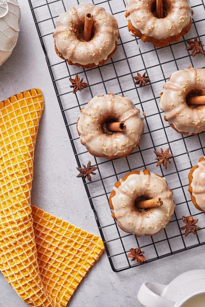 Overhead view of mini pumpkin bundt cakes on a wire rack with cinnamon sticks, orange kitchen cloth on a marble countertop.