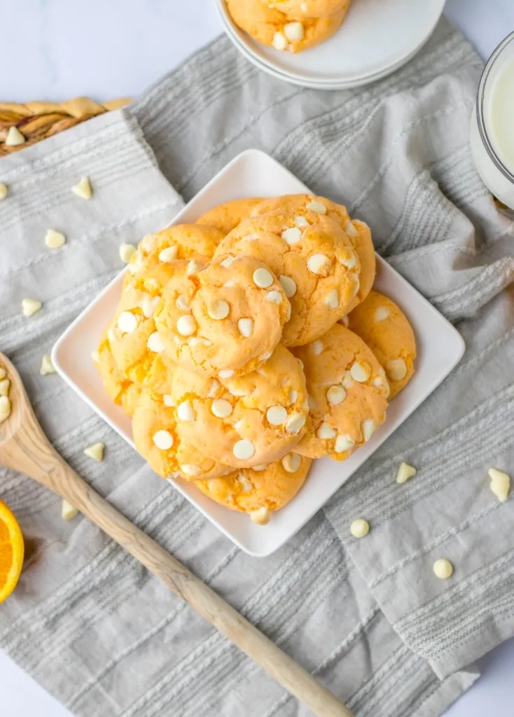 Overhead view of stacked orange creamsicle cookies on a white plate, wooden spoon, grey kitchen cloth, on a marble countertop.