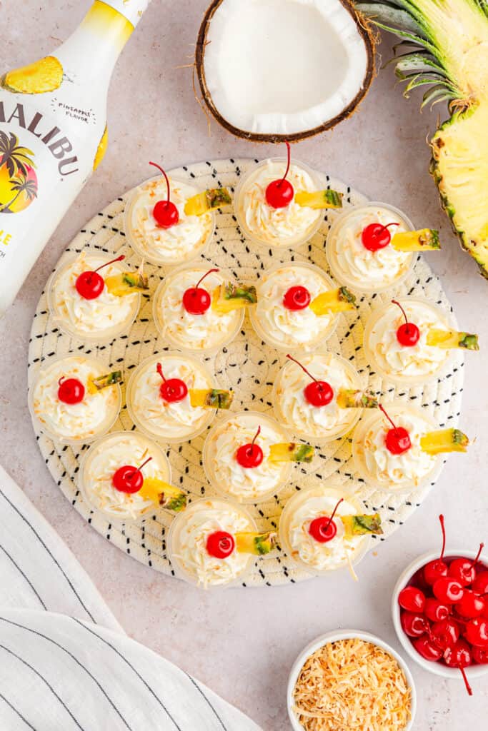 Overhead view of Pina Colada Jello shots on a decorative mat surrounded by the recipe ingredients, striped linen cloth on a white countertop.