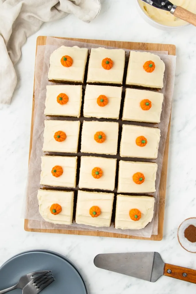 Sliced pumpkin cake with cream cheese frosting on a wooden board, metal cake server, serving plates with forks on a marble countertop.