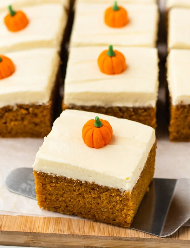 Close up of slices of pumpkin cake with cream cheese frosting on a wooden board with metal server.