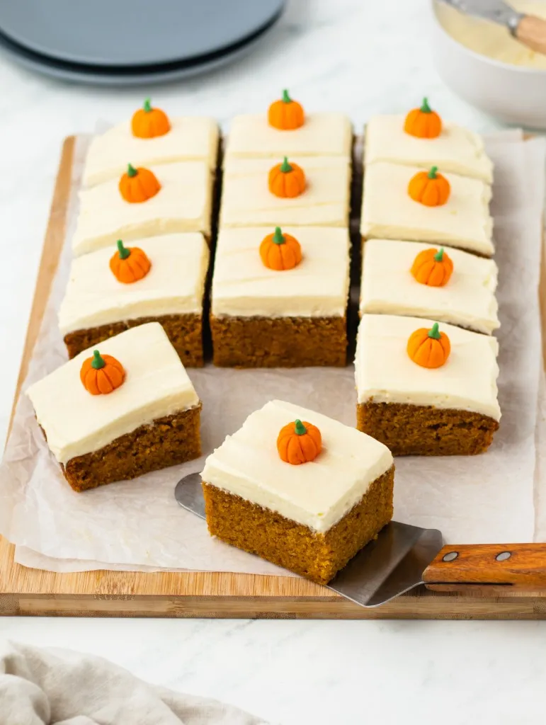 Close up of sliced pumpkin cake with cream cheese frosting, piece of cake on a metal server, serving plates, cream linen cloth on a white marble countertop.
