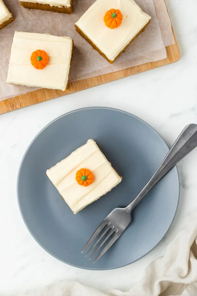 Overhead view of a slice of pumpkin cake with cream cheese frosting on a serving plate with metal fork, slices of cake on a wooden board, cream kitchen linen on a white marble countertop.