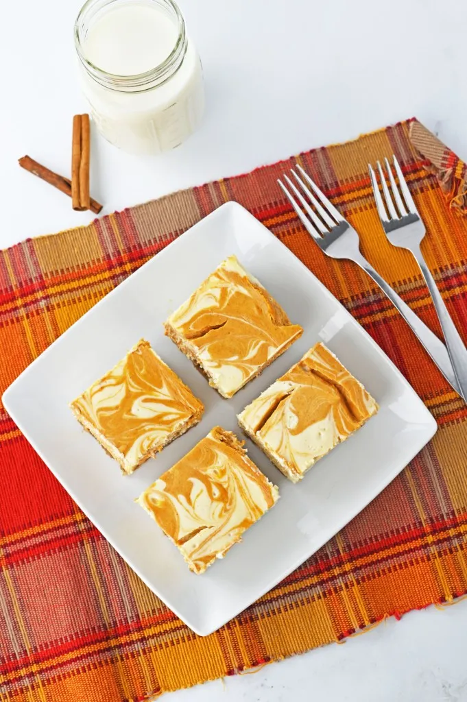 Overhead view of pumpkin cheesecake bars on a white plate, two metal forks, glass of milk, cinnamon sticks, orange checkered cloth.