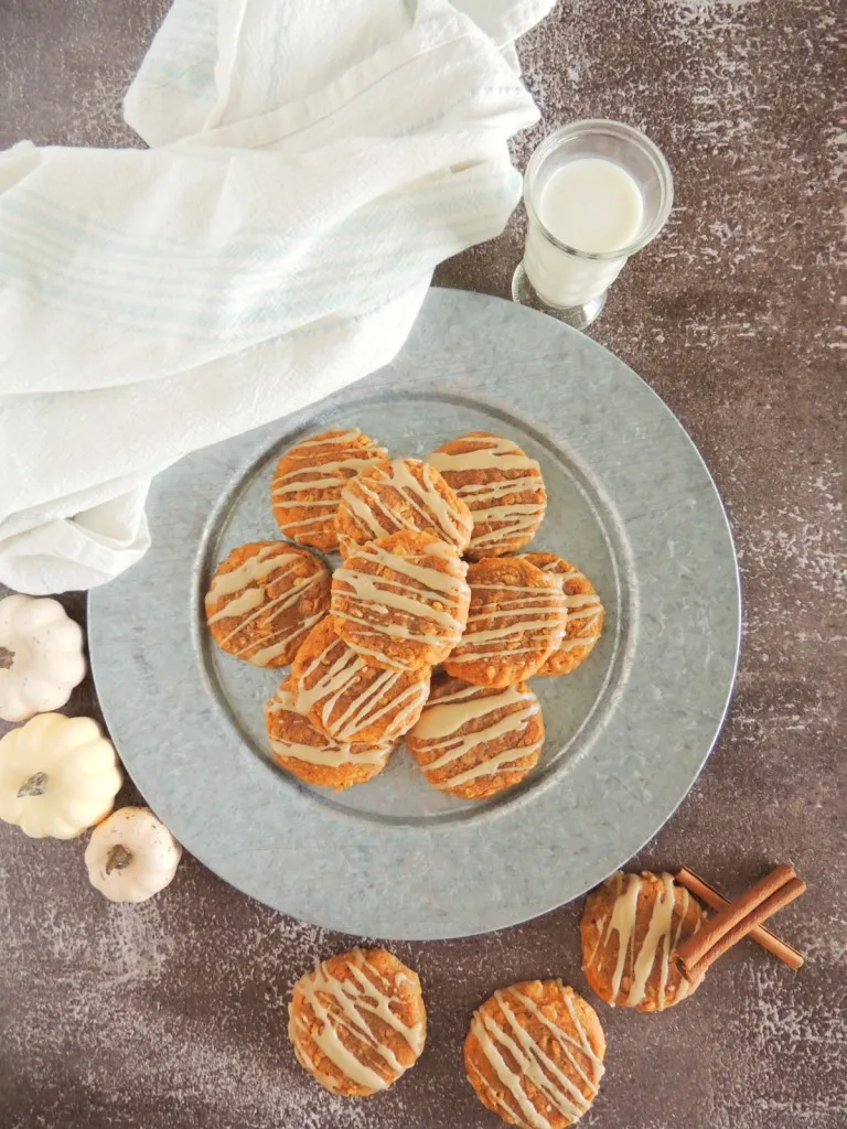 Pumpkin oatmeal cookies on a grey plate, additional cookies scattered around the plate, glass of milk, white kitchen towel on a wooden surface.