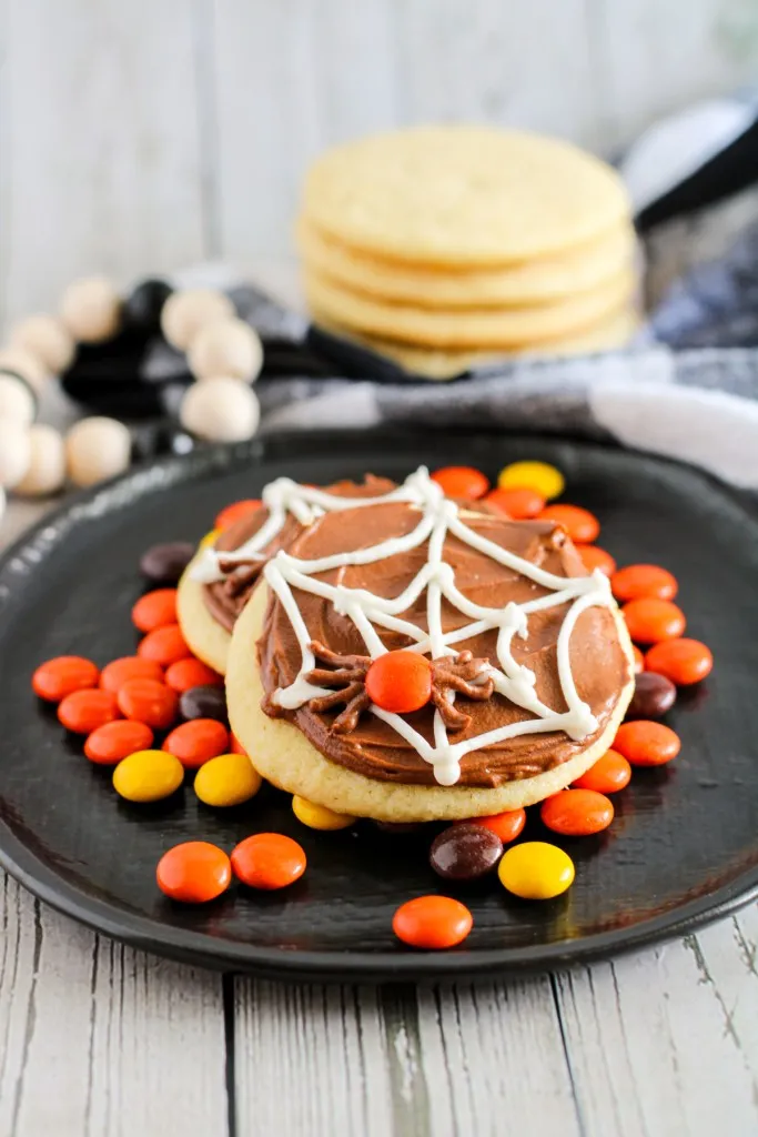 Easy spider web sugar cookies on a black plate with Reese's Pieces, plain sugar cookies in the background.