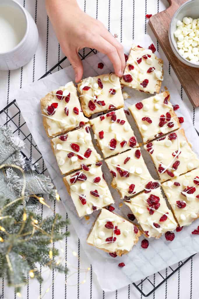 Hand reaching for a cranberry bliss bar, remaining cranberry bliss bars on parchment paper on a wire cooling rack.