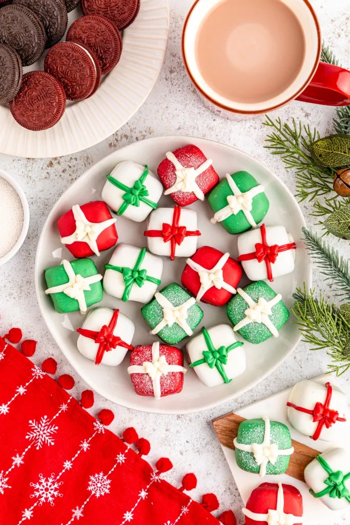 Overhead view of a plate of Christmas Oreo presents, plate of Oreo cookies, mug of hot chocolate, Christmas-themed table decorations.