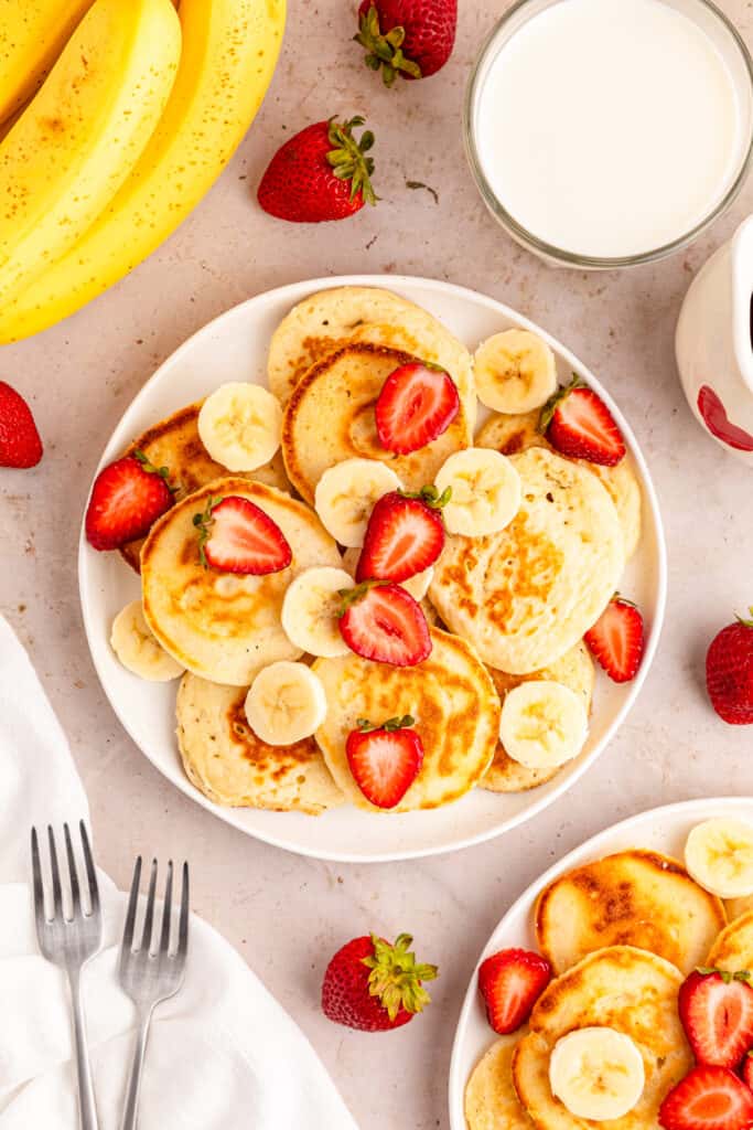 a close up view of mini pancakes garnished with sliced bananas and strawberries and a glass of milk